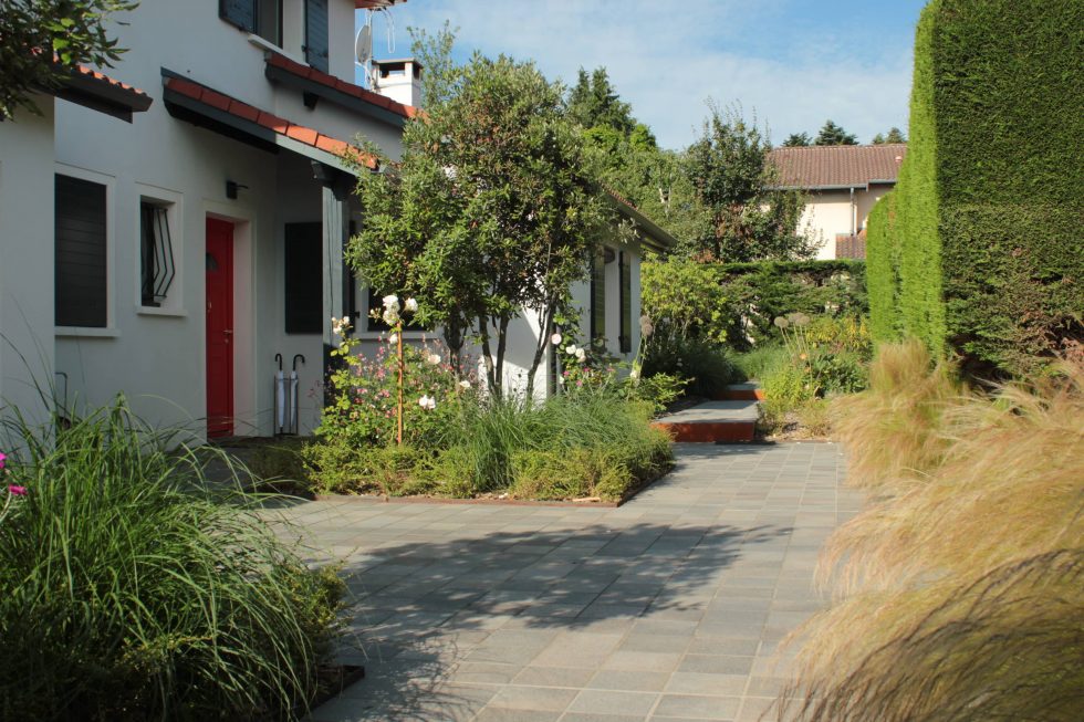 Photo de la cour et accès de cette maison à Saint Didier au Mont d'Or : carreaux petits formats aux teintes nordiques, mis en valeurs par deux grands carrés plantés. Cet aménagement offre une arrivée accueillante et vivante, à pied ou en voiture.