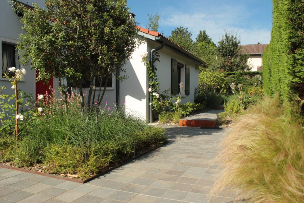 Photo de la cour et accès de cette maison à Saint Didier au Mont d'Or : carreaux petits formats aux teintes nordiques, mis en valeurs par deux grands carrés plantés. Cet aménagement offre une arrivée accueillante et vivante, à pied ou en voiture.