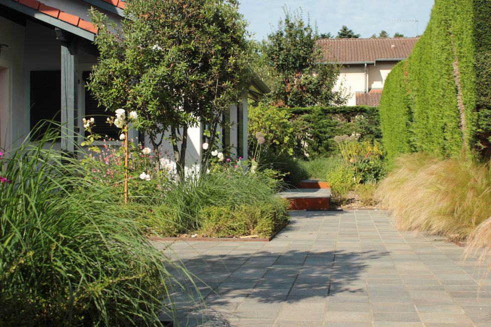 photo du réaménagement d'un jardin à Saint Didier au Mont d'Or. Création de carrés plantés : des plantations luxuriantes dans des lignes droites. Nous avons profité des légères différences de niveau pour créer un allée bordée de végétation et ponctuée de quelques marches. Photo de la cour et accès de cette maison à Saint Didier au Mont d'Or : carreaux petits formats aux teintes nordiques, mis en valeurs par deux grands carrés plantés. Cet aménagement offre une arrivée accueillante et vivante, à pied ou en voiture.